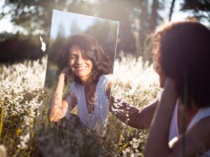 Een stralende vrouw houdt een spiegel vast in de natuur, terwijl de zon haar gezicht verlicht. Ze glimlacht met zelfvertrouwen en houdt van zichzelf.